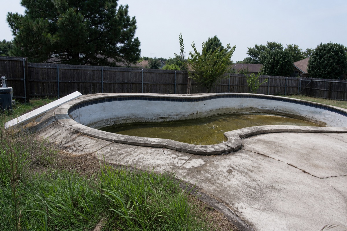 Empty freeform pool with cracked decking and stagnant water, typical of a remodel-or-rebuild decision.